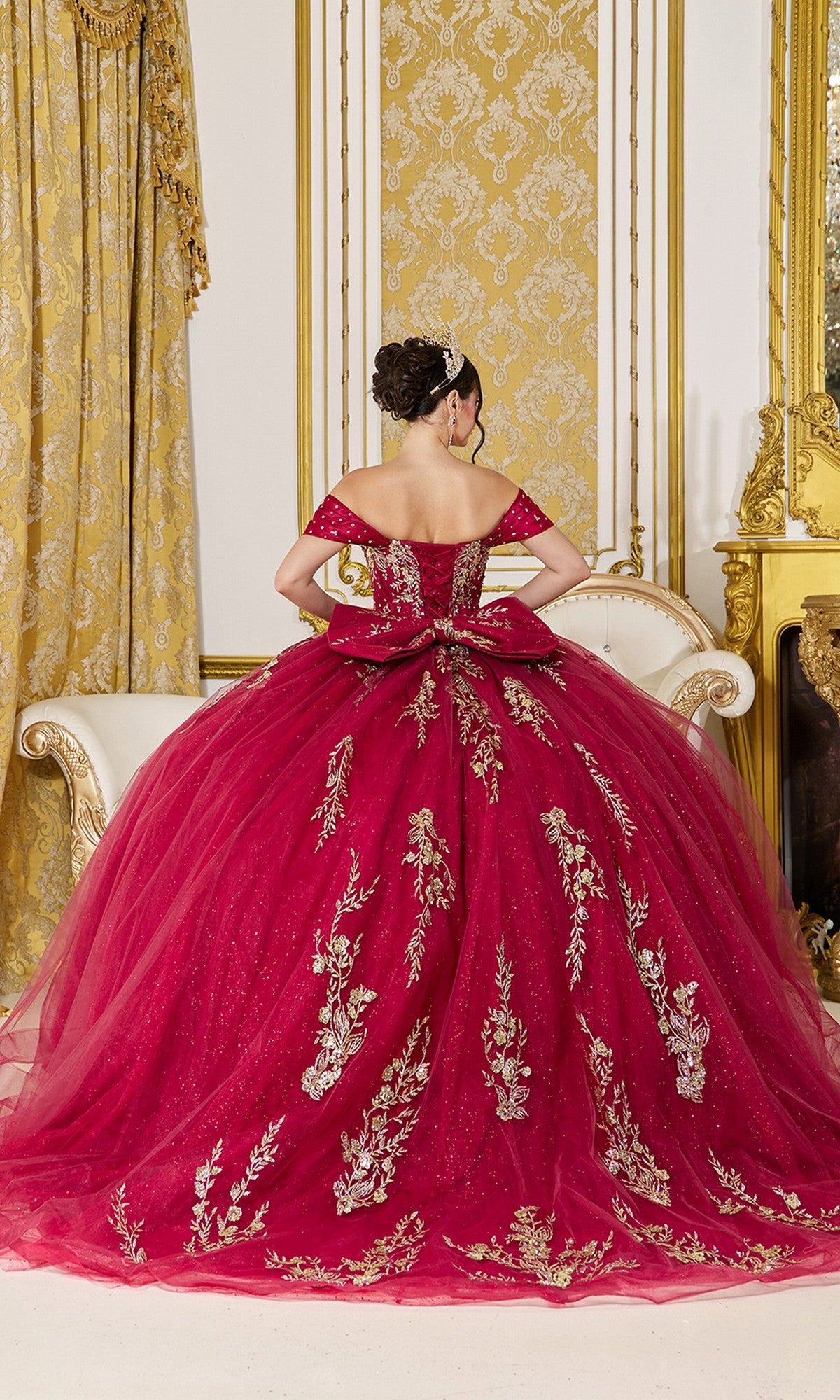 A woman stands with her back to the camera in an elegant, gold-accented room, wearing the Quinceanera Dress 5067 By Dancing Queen—a burgundy off-the-shoulder ball gown with gold floral embroidery and a large bow.